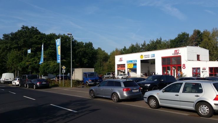 Eine Straße mit mehreren am Straßenrand geparkten Autos, ein Gebäude mit verschiedenen Schildern und ein paar Bäumen im Hintergrund.