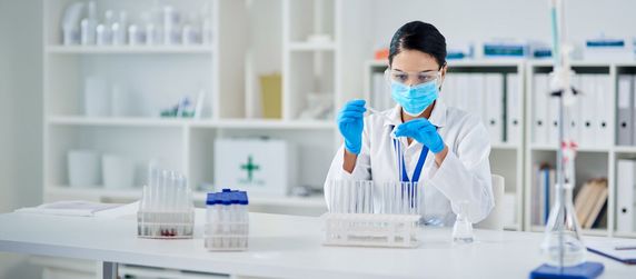 Eine Frau in einem Laborkittel und blauen Handschuhen arbeitet mit einem Reagenzglas.