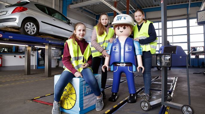 Vier Personen in einer Werkstatt, in der Mitte eine große Playmobil-Figur, die als TUEV Hessen Mechaniker verkleidet ist.