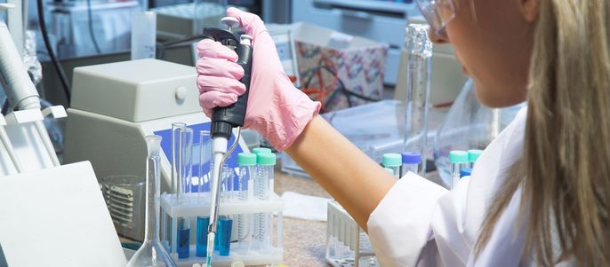 Eine Laborumgebung, in der eine Person mit Schutzausrüstung eine Pipette benutzt, um eine Flüssigkeit in ein Reagenzglas zu füllen.