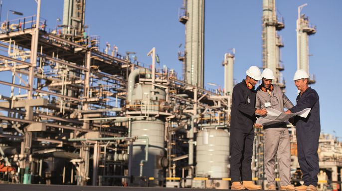 Eine Gruppe von Arbeitern auf einem Industriegelände, die einen Bauplan begutachten. Die Baustelle besteht aus hohen Strukturen, Rohren und Geräten. Die Arbeiter tragen Schutzhelme und Schutzkleidung.