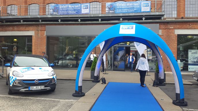 Eine Veranstaltung im Freien mit einem blauen Auto, einem blauen Teppich und einem blauen Torbogen. Es gibt Transparente und Schilder, und eine Person geht auf den Eingang zu.