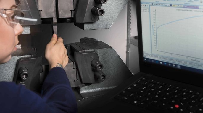 Eine Person mit Schutzbrille, die eine Maschine einstellt, während im Hintergrund eine Grafik auf einem Laptop-Bildschirm zu sehen ist.