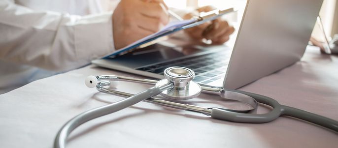 Eine medizinische Fachkraft, vermutlich ein Arzt, benutzt einen Laptop und hält ein Klemmbrett. Auf dem Tisch neben dem Laptop befindet sich ein Stethoskop.